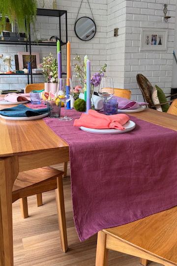 A styled dining table in a bright room with a wooden table and chairs, featuring a prominent magenta table runner, coral napkins, colorful candles, and floral centerpieces against a white brick wall background.