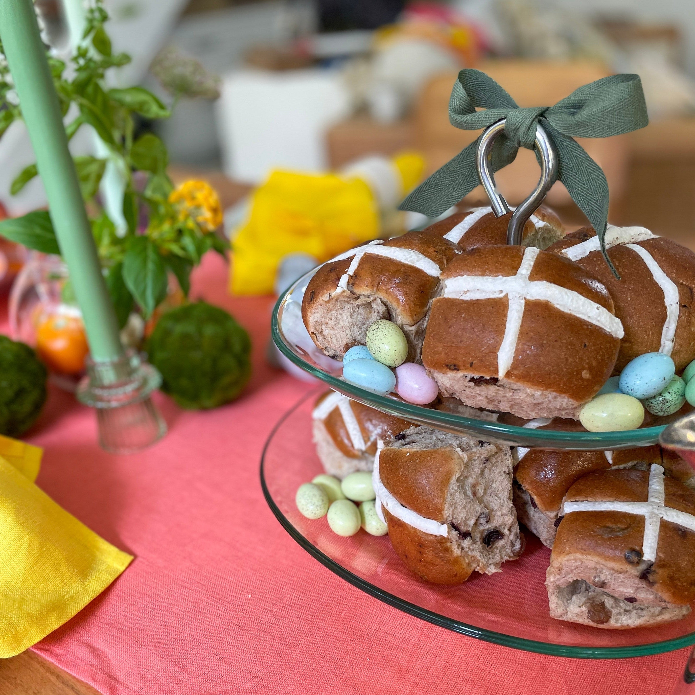 Two-tiered glass stand with hot cross buns and Easter eggs on a pink tablecloth.